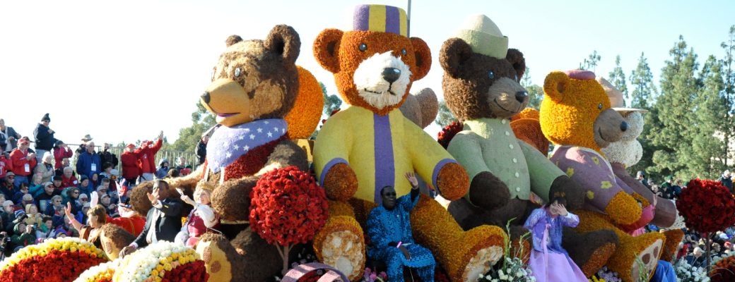 Colorful float with large teddy bear figures made of flowers in a parade, with spectators watching in the background.