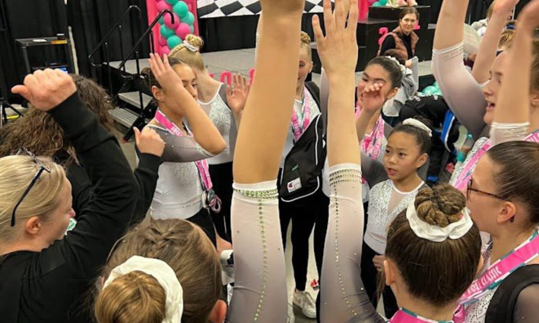 Young gymnasts in leotards giving a team high-five backstage, with a coach joining in, expressing excitement and teamwork.
